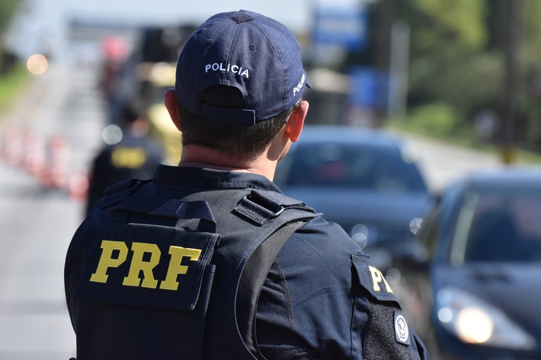 Agente da Polícia Rodoviária Federal de costas, com identificação PRF no uniforme, durante fiscalização de veículos em uma rodovia.