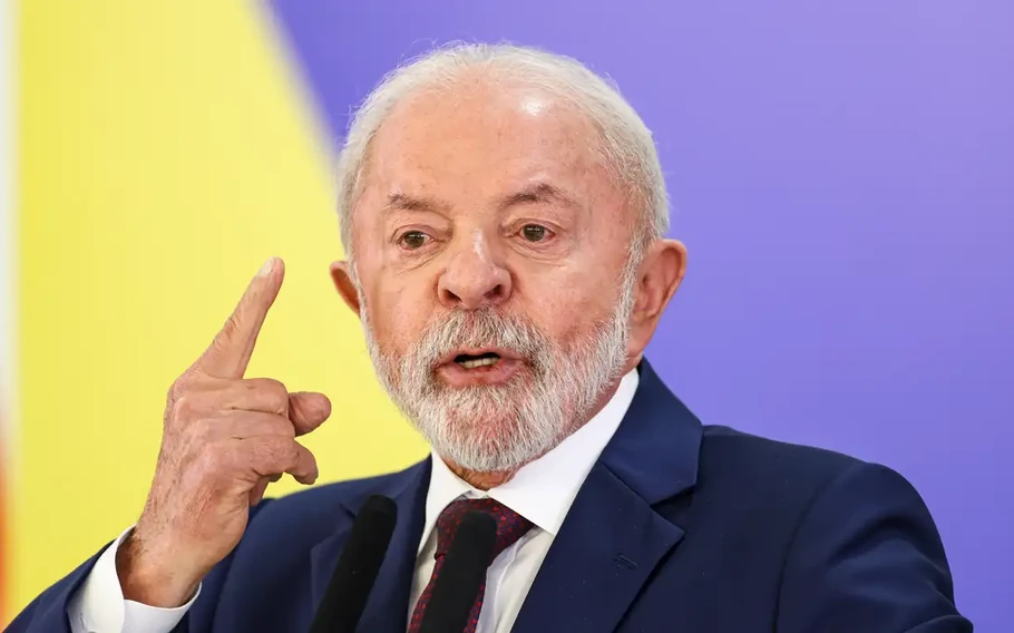 Presidente Lula, de terno azul-escuro, fala diante de microfones com o dedo indicador levantado, sobre fundo amarelo e lilás.