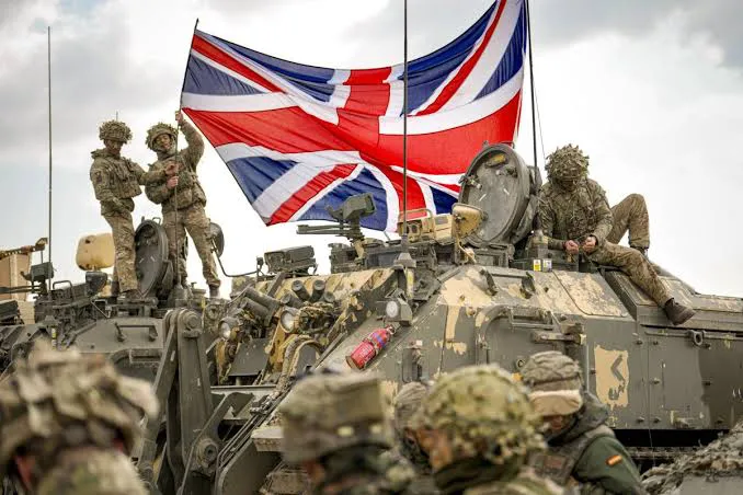 Militares britânicos sobre veículos blindados, com uma grande bandeira do Reino Unido ao fundo.