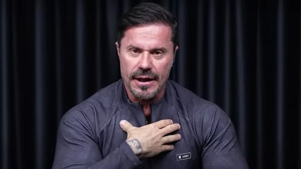 Renato Cariani, de camiseta esportiva cinza, fala diante de um fundo preto com uma das mãos apoiada sobre o peito.
