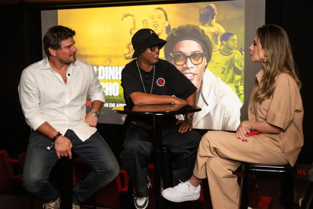 Ronaldinho Gaúcho, de camiseta e chapéu pretos e óculos escuros, conversa com um homem de camisa branca e uma mulher de roupa bege diante de um painel.