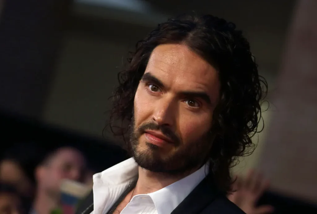 Russell Brand, ator e comediante britânico, com cabelos compridos e barba, usando paletó escuro e camisa branca em fotografia de arquivo.