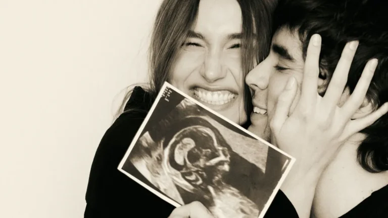 Sasha Meneghel e João Lucas aparecem sorridentes, abraçados e segurando uma imagem de ultrassom diante da câmera.