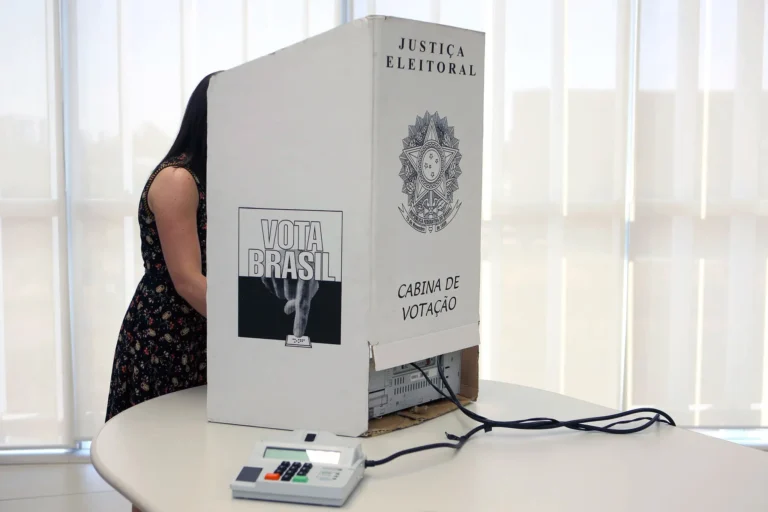 Eleitora atrás de uma cabine de votação da Justiça Eleitoral, com urna eletrônica sobre a mesa.