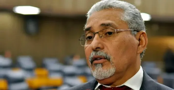 Senival Moura, de cabelos grisalhos, óculos, bigode e cavanhaque, veste paletó escuro, camisa branca e gravata vinho, com cadeiras desfocadas ao fundo.