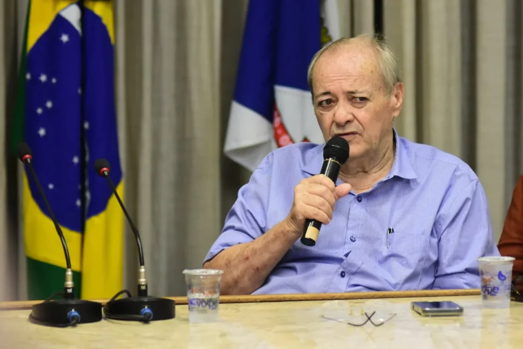 Prefeito Silvio Mendes, de camisa azul-clara, sentado à mesa e segurando um microfone, com a bandeira do Brasil e outra bandeira ao fundo.