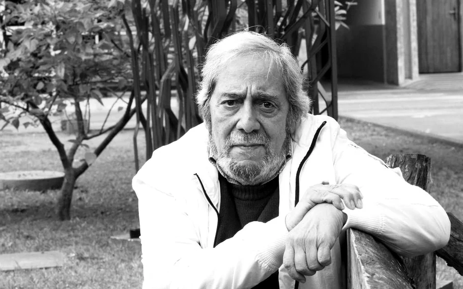 Retrato em preto e branco de Solano Ribeiro, com cabelos e barba grisalhos, usando casaco claro e apoiando os braços em uma estrutura de madeira, em área ajardinada.