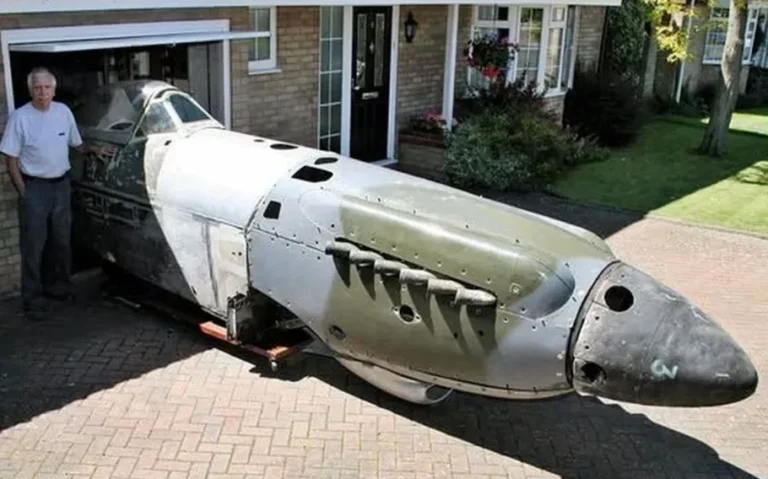 Fuselagem de um avião militar sem asas posicionada diante da garagem de uma casa, com um homem de camiseta clara ao lado.