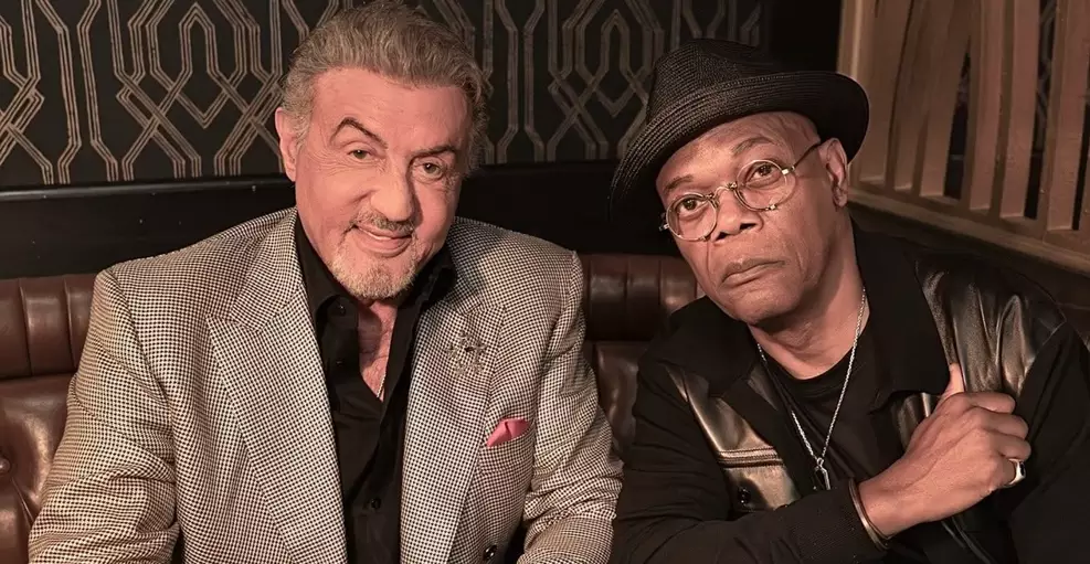 Sylvester Stallone, de paletó xadrez, posa ao lado de Samuel L. Jackson, que usa chapéu preto, óculos e colete de couro, em ambiente interno.