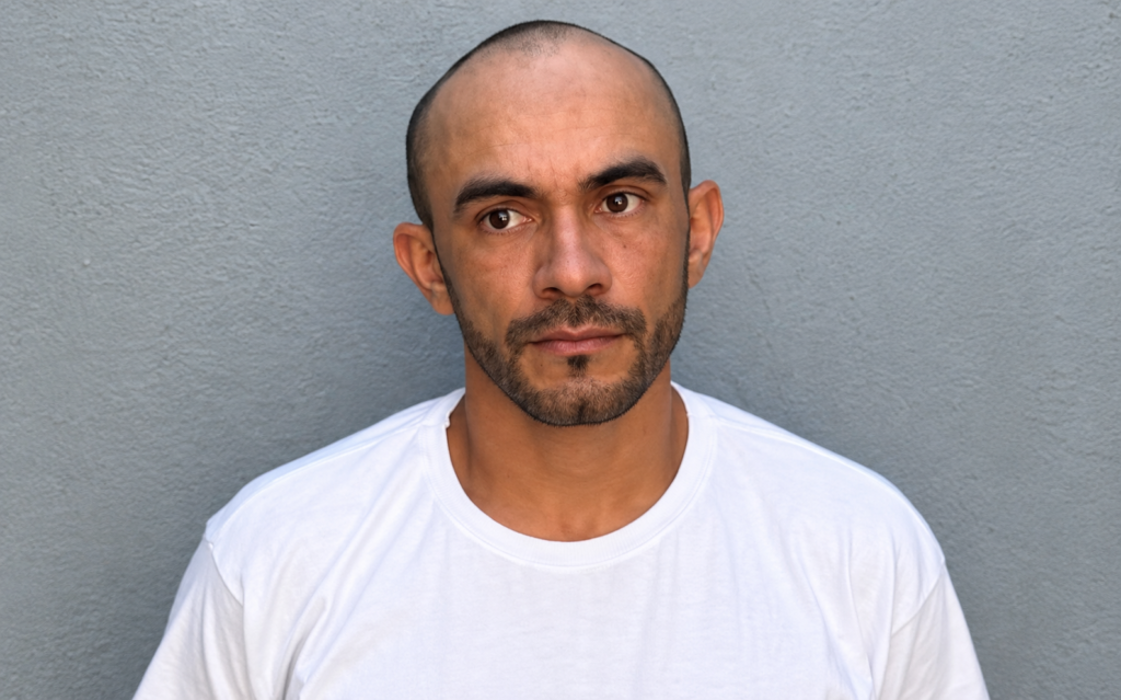 Retrato de homem de cabeça raspada, barba curta e camiseta branca diante de parede cinza.