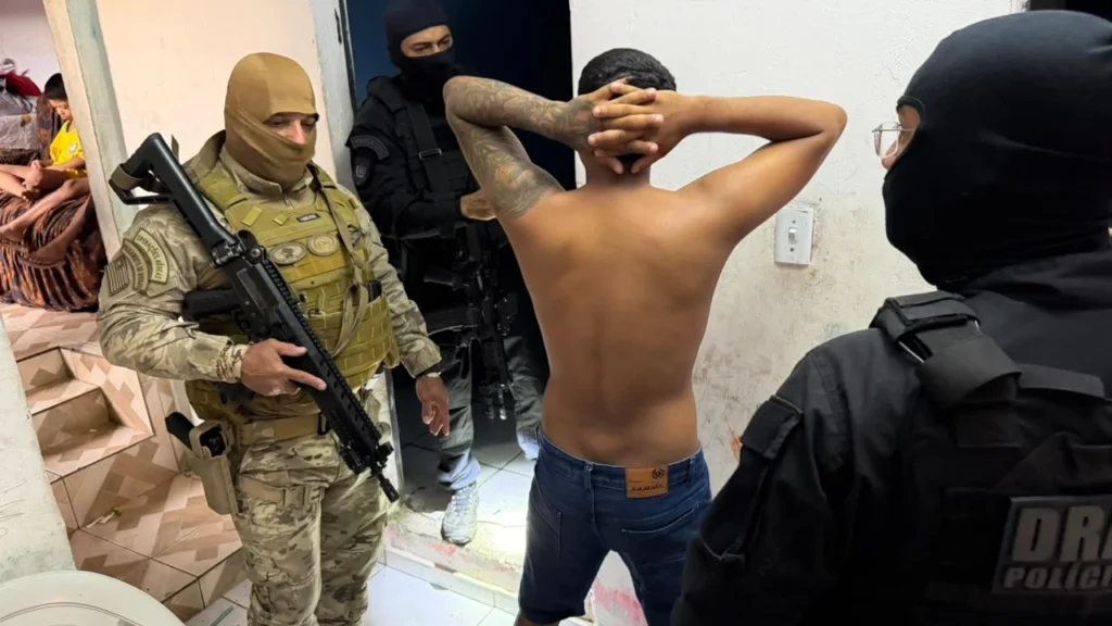Homem sem camisa, de costas e com as mãos sobre a cabeça, cercado por policiais encapuzados durante uma abordagem.