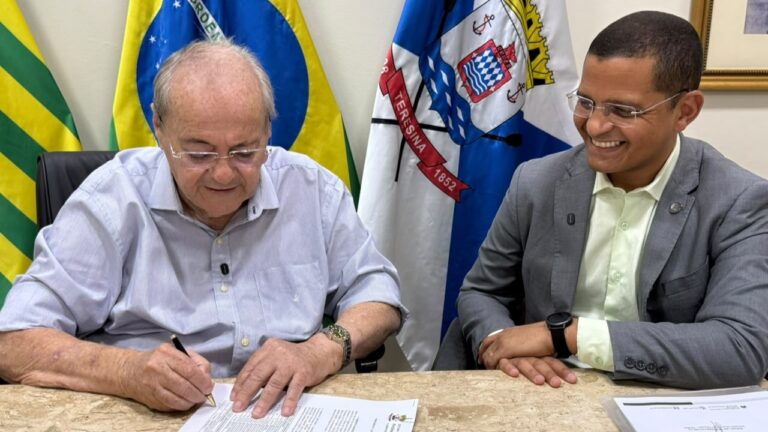 Prefeito Silvio Mendes assinando documento de convocação de professores, ao lado do secretário de Educação Ismael Silva, com bandeiras ao fundo.