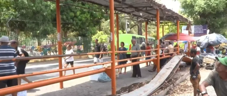 Parada de ônibus com pilares e corrimãos alaranjados, parte da cobertura caída na lateral e pessoas próximas à estrutura danificada.