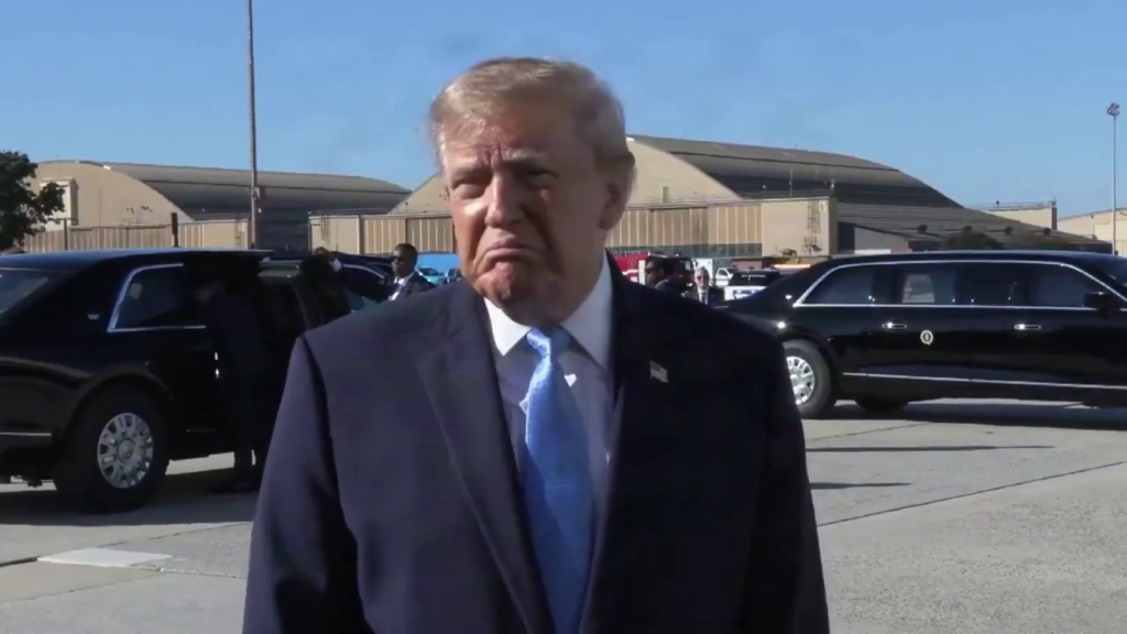 Donald Trump, de terno escuro e gravata azul, fala à imprensa em área externa, diante de veículos oficiais.