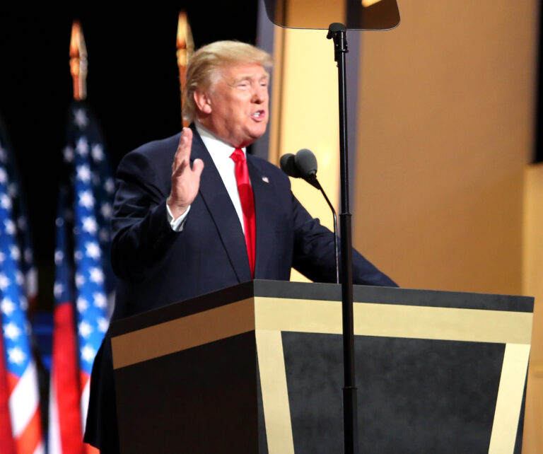 Donald Trump, de terno escuro e gravata vermelha, discursa ao microfone com uma mão levantada e bandeiras dos Estados Unidos ao fundo.