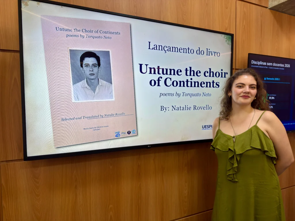 Natalie Rovello, de vestido verde, ao lado de uma tela que exibe a capa da coletânea de poemas de Torquato Neto em inglês, com o retrato do poeta.