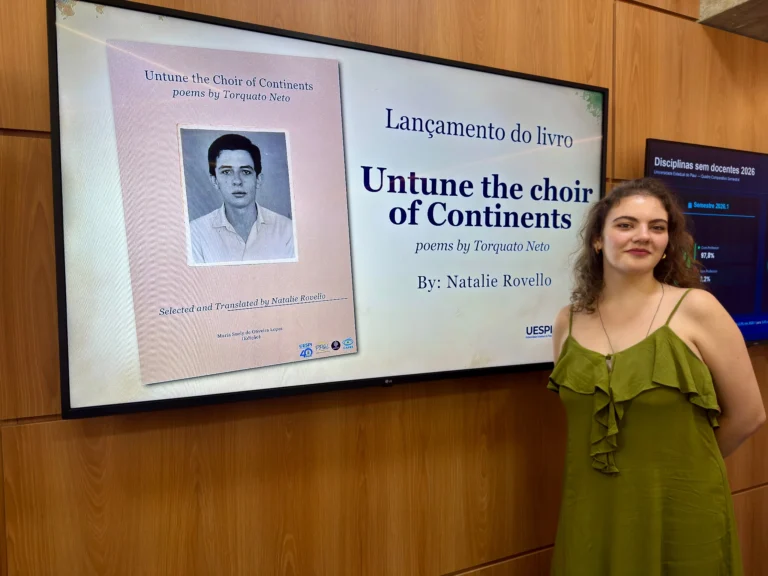 Natalie Rovello, de vestido verde, ao lado de uma tela que exibe a capa da coletânea de poemas de Torquato Neto em inglês, com o retrato do poeta.