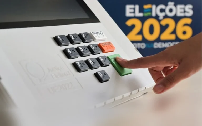 Dedo pressiona a tecla verde Confirma de uma urna eletrônica, com a identificação Eleições 2026 ao fundo.