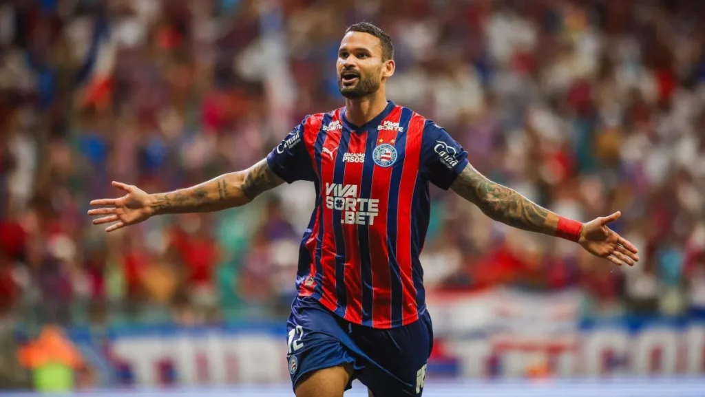 Willian José, com uniforme azul e vermelho do Bahia, comemora de braços abertos em campo, diante de arquibancadas desfocadas.