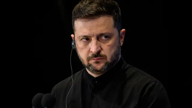 Volodymyr Zelensky, de camisa preta e fone de ouvido, olha para o lado diante de microfones, com fundo escuro.
